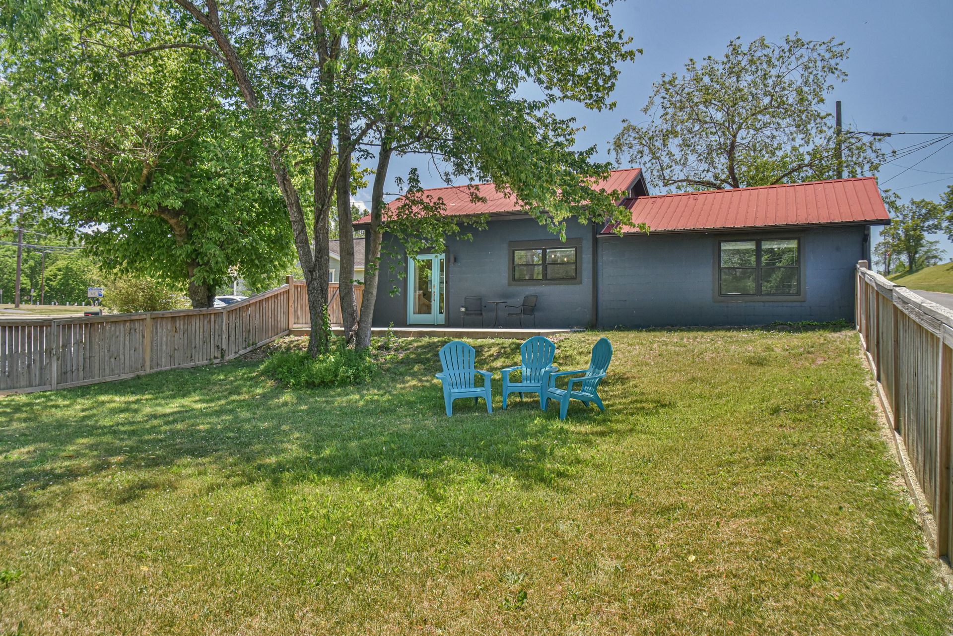 Backyard with green lawn, blue chairs, and a small house with a red roof, framed by wooden fences and trees