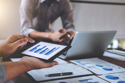 Two people reviewing business data on a tablet, with charts and graphs on paper spread across a desk.