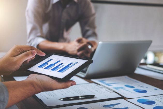 Two people reviewing business data on a tablet, with charts and graphs on paper spread across a desk.