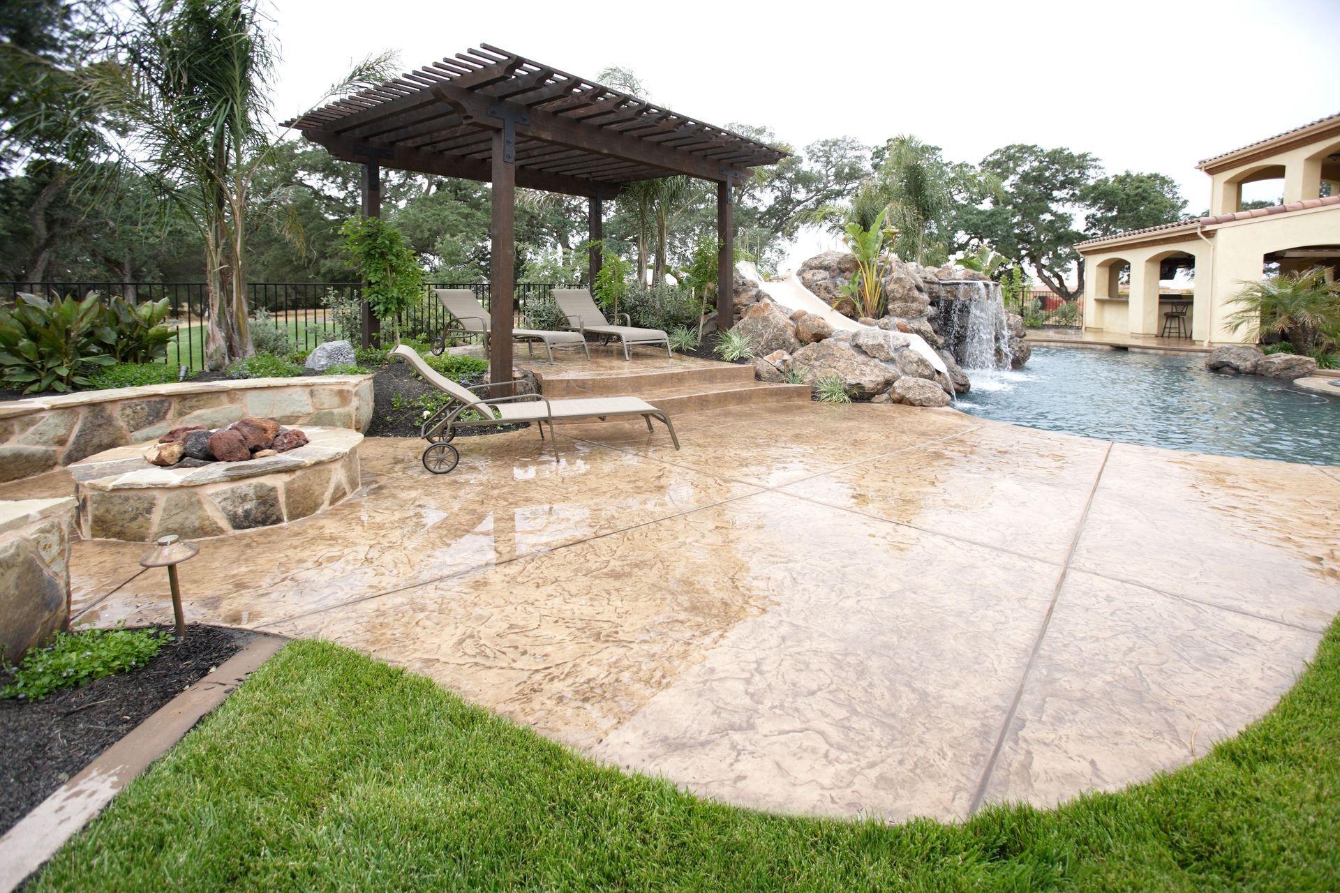 Backyard patio with gazebo, stone waterfall pool, and wet tiled deck beside a green lawn