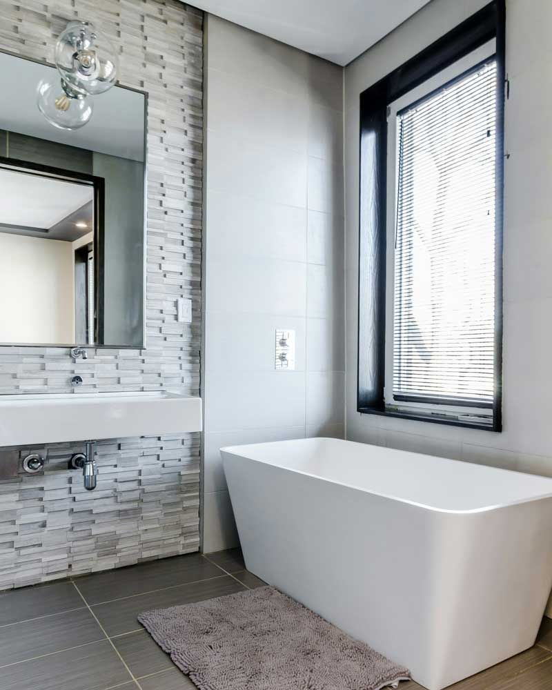 Modern bathroom with a freestanding white tub, gray tile floor, mosaic wall, and tall window blinds