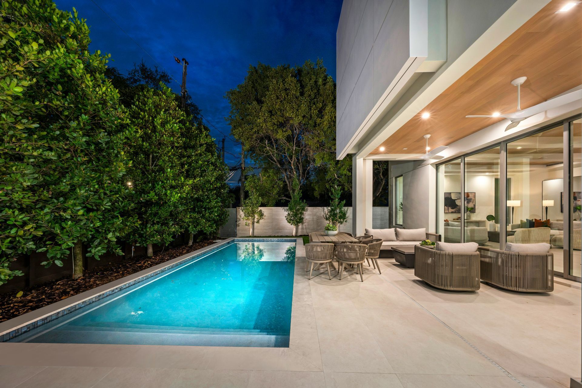 Modern patio with illuminated pool, lounge chairs, and a glass-walled house at dusk