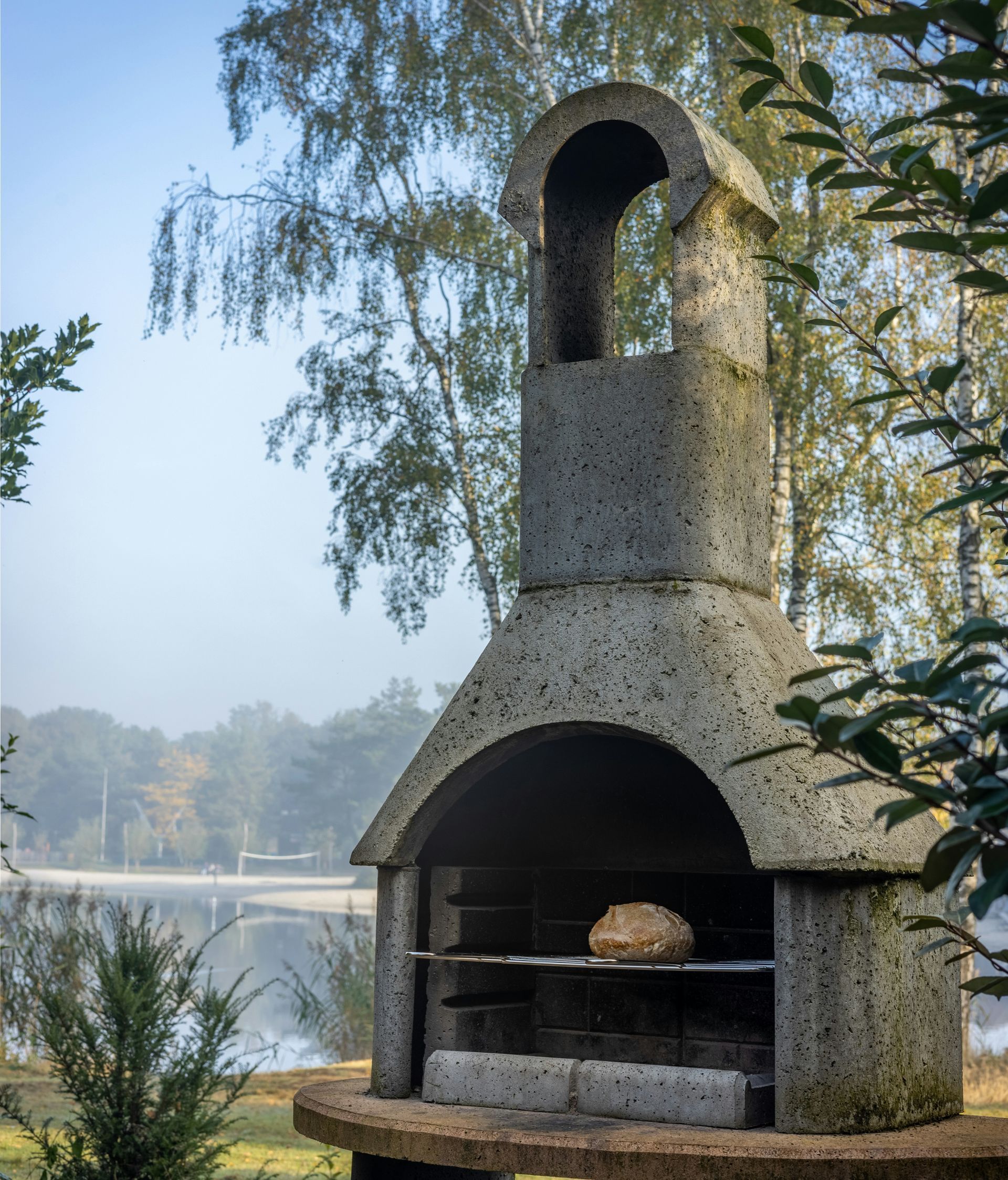 Stone outdoor fireplace by a lake, with arched openings and trees in the background