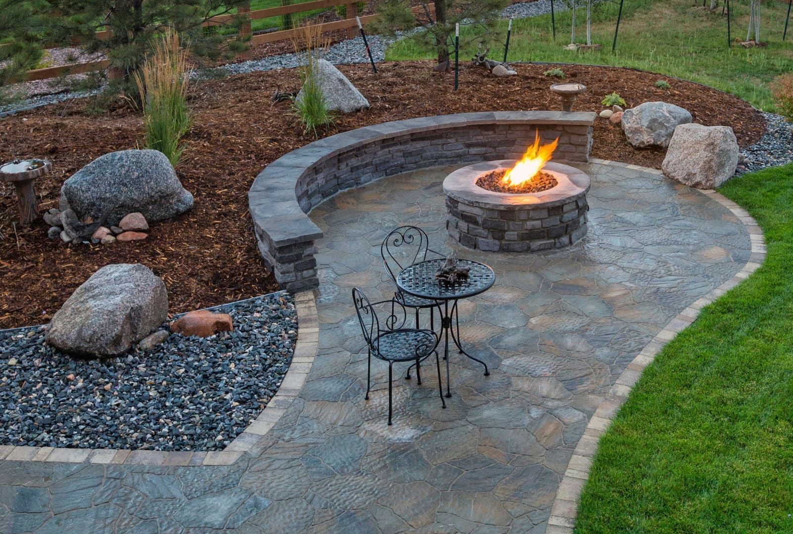 Stone patio with a lit fire pit, metal chairs, and landscaped rock garden in a backyard