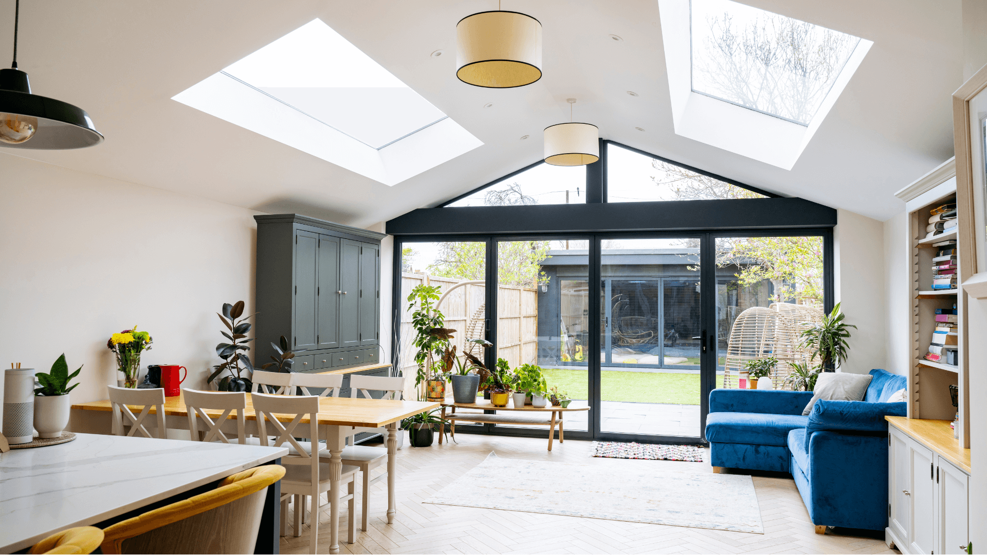 Bright modern living room with skylights, large windows, blue sofa, dining table, plants, and garden view