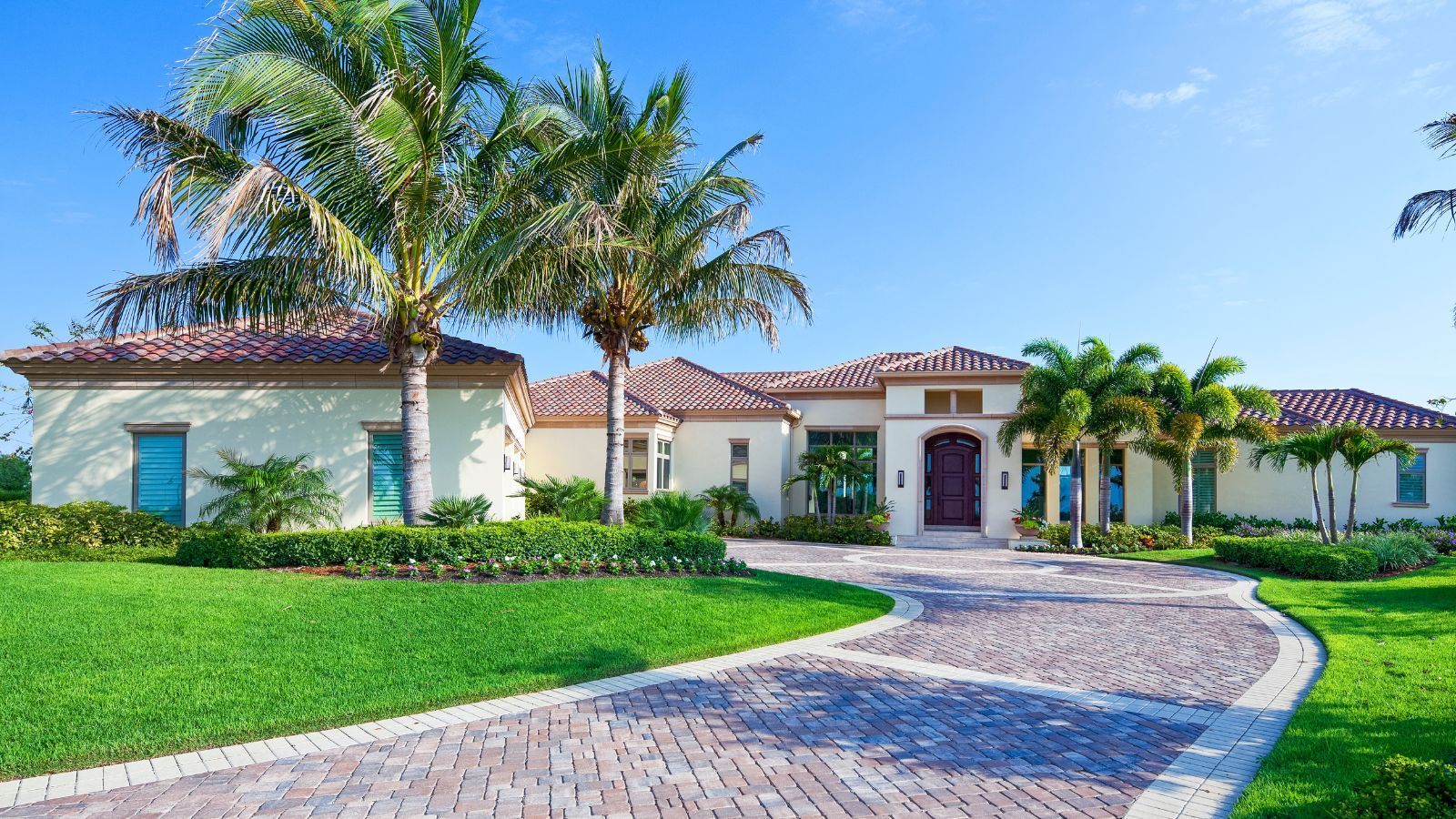 Luxury single-story house with a tiled roof, palm trees, and a curved brick driveway under a blue sky