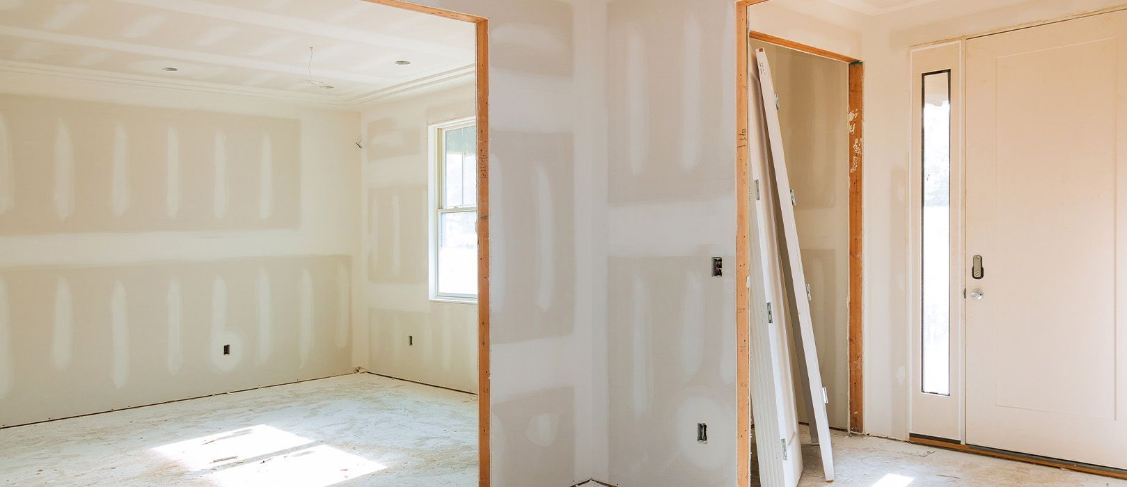 Empty drywall room under construction with white walls, exposed studs, and stacked boards by a window.