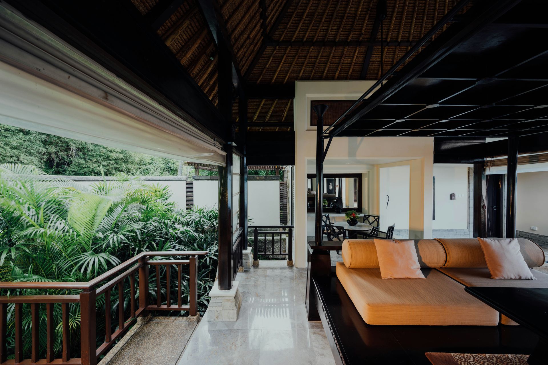 Covered tropical veranda with wooden lounge, orange cushions, and lush greenery outside