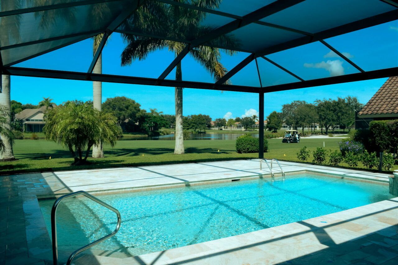 Covered backyard swimming pool with blue water, patio, and palm trees under a shaded canopy.