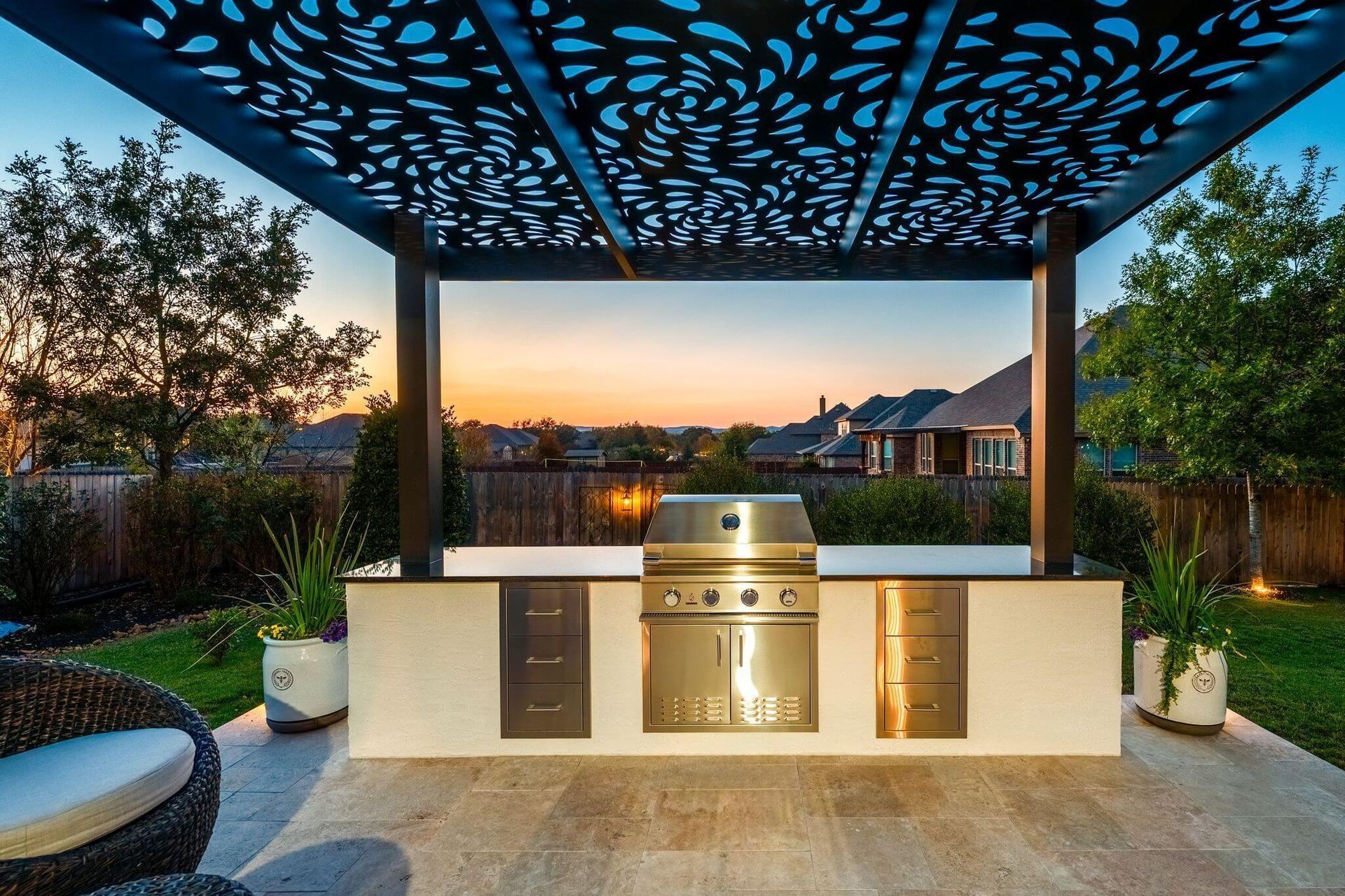 Outdoor patio with a built-in grill under a decorative pergola at sunset, overlooking a backyard.