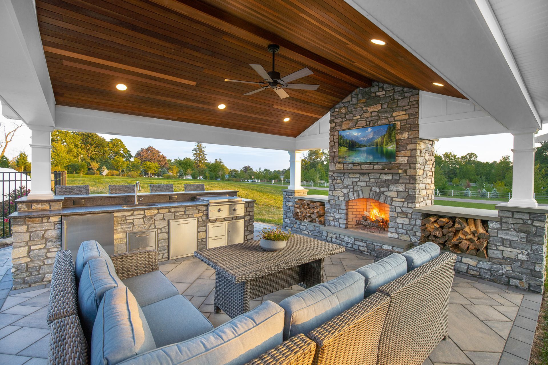 Outdoor covered patio with stone fireplace, built-in grill, and gray sectional seating under a wood ceiling