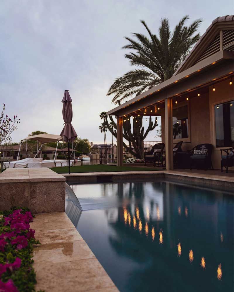 Resort pool beside a patio at dusk, with palm trees, lounge chairs, and warm string lights reflected in water