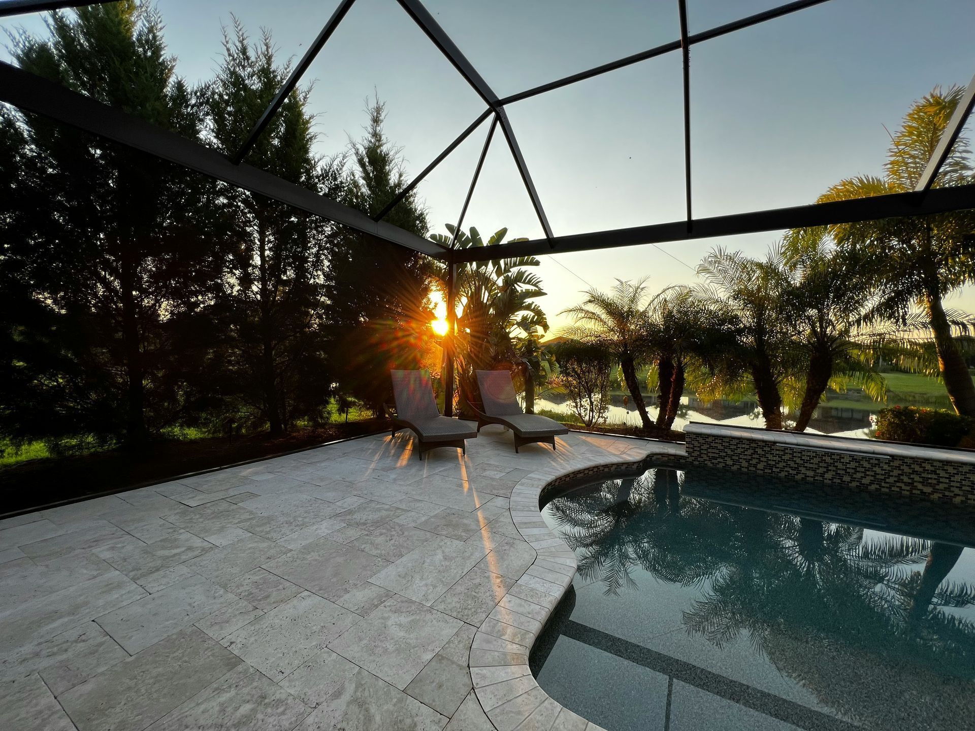 Sunset view of a screened patio with lounge chairs beside a swimming pool and palm trees