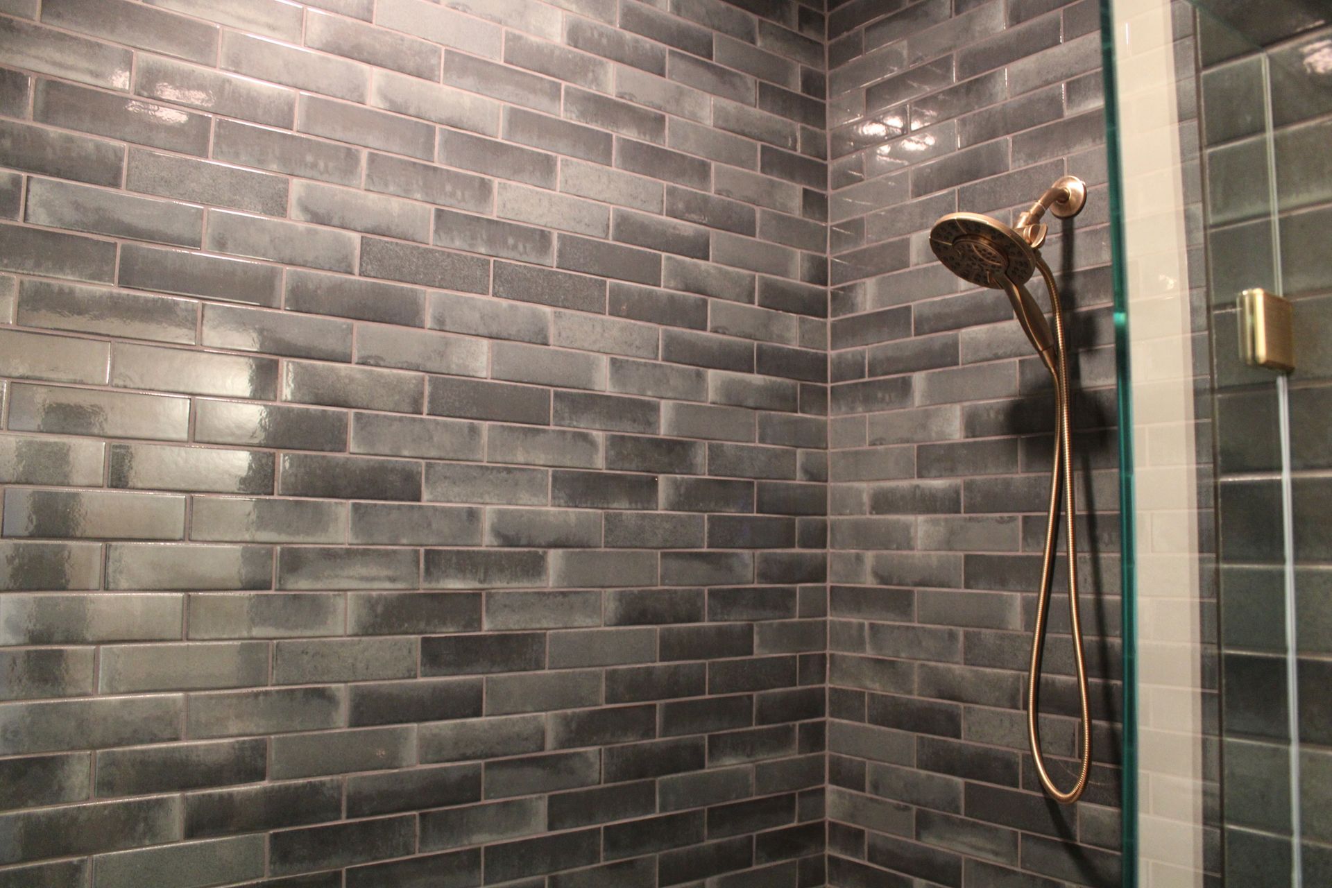 Gray tiled shower with a bronze handheld showerhead and glass door