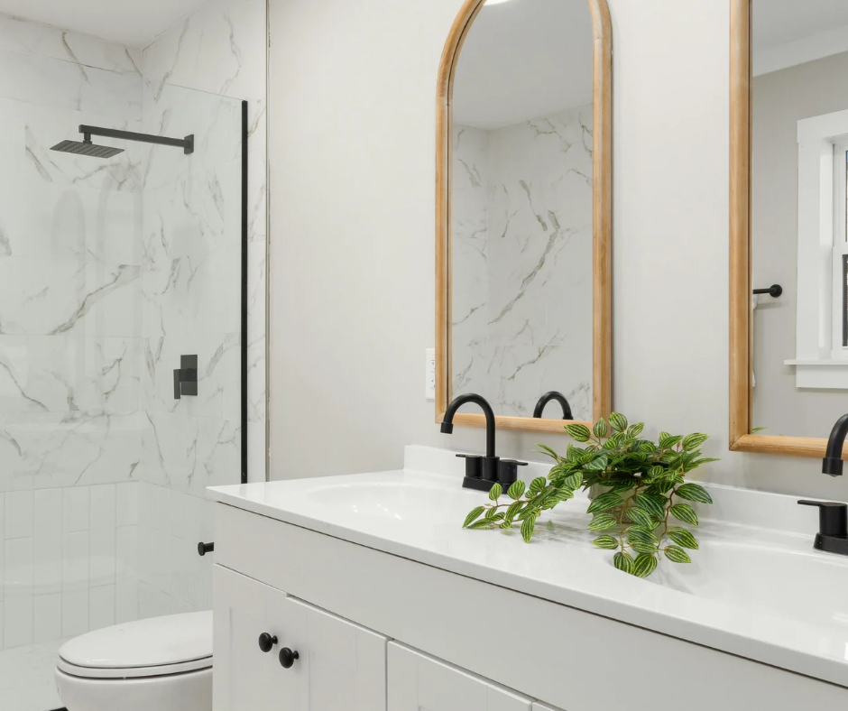 Modern white bathroom with marble shower, black fixtures, gold-framed mirror, and a vanity with green plant
