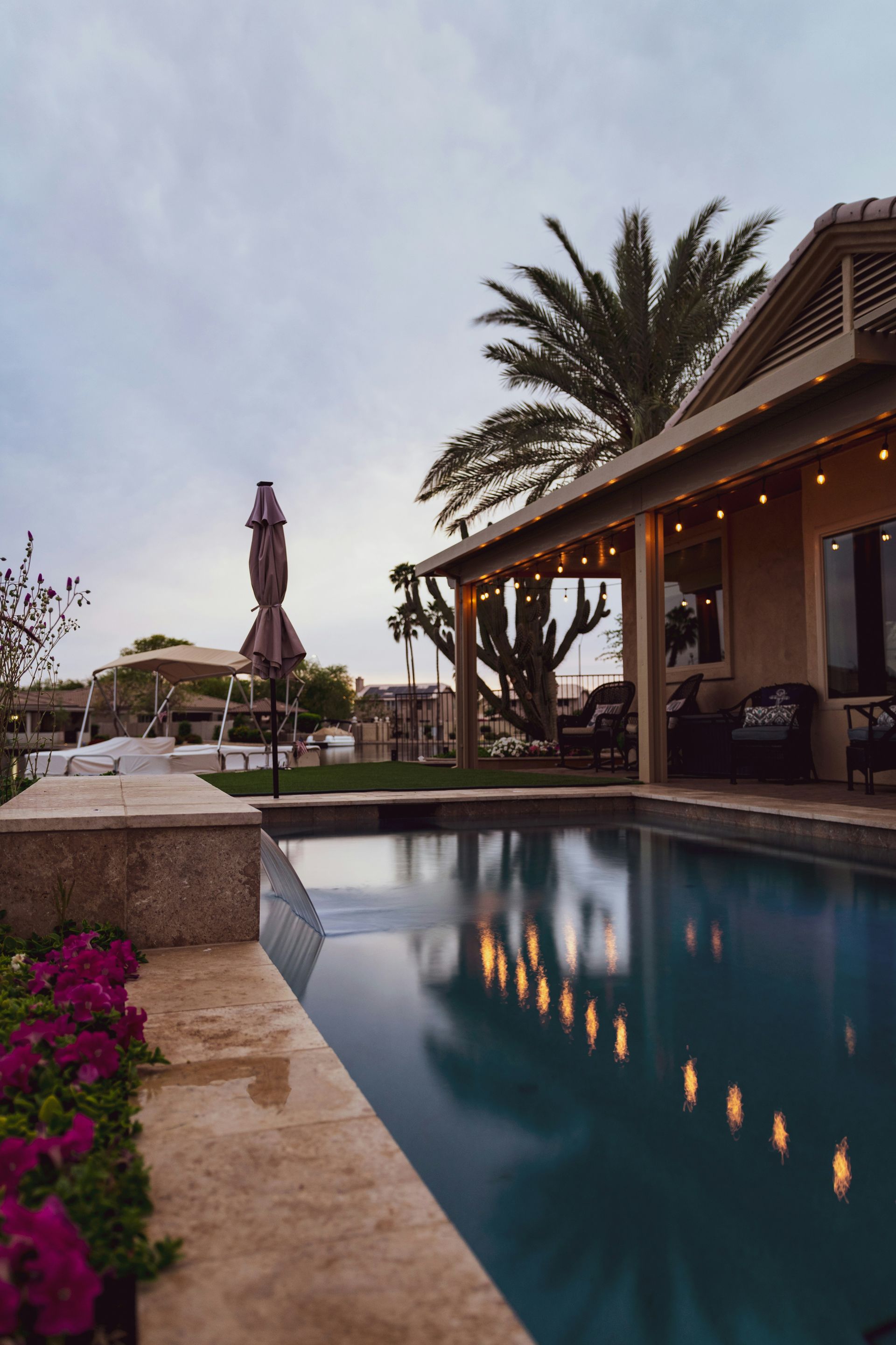 Resort pool at dusk with palm trees, lit patio, and a closed umbrella by the water