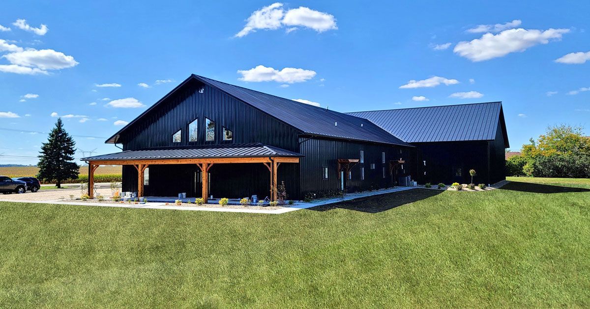Black barn-style building with a porch, on a grassy field under a blue sky with clouds.