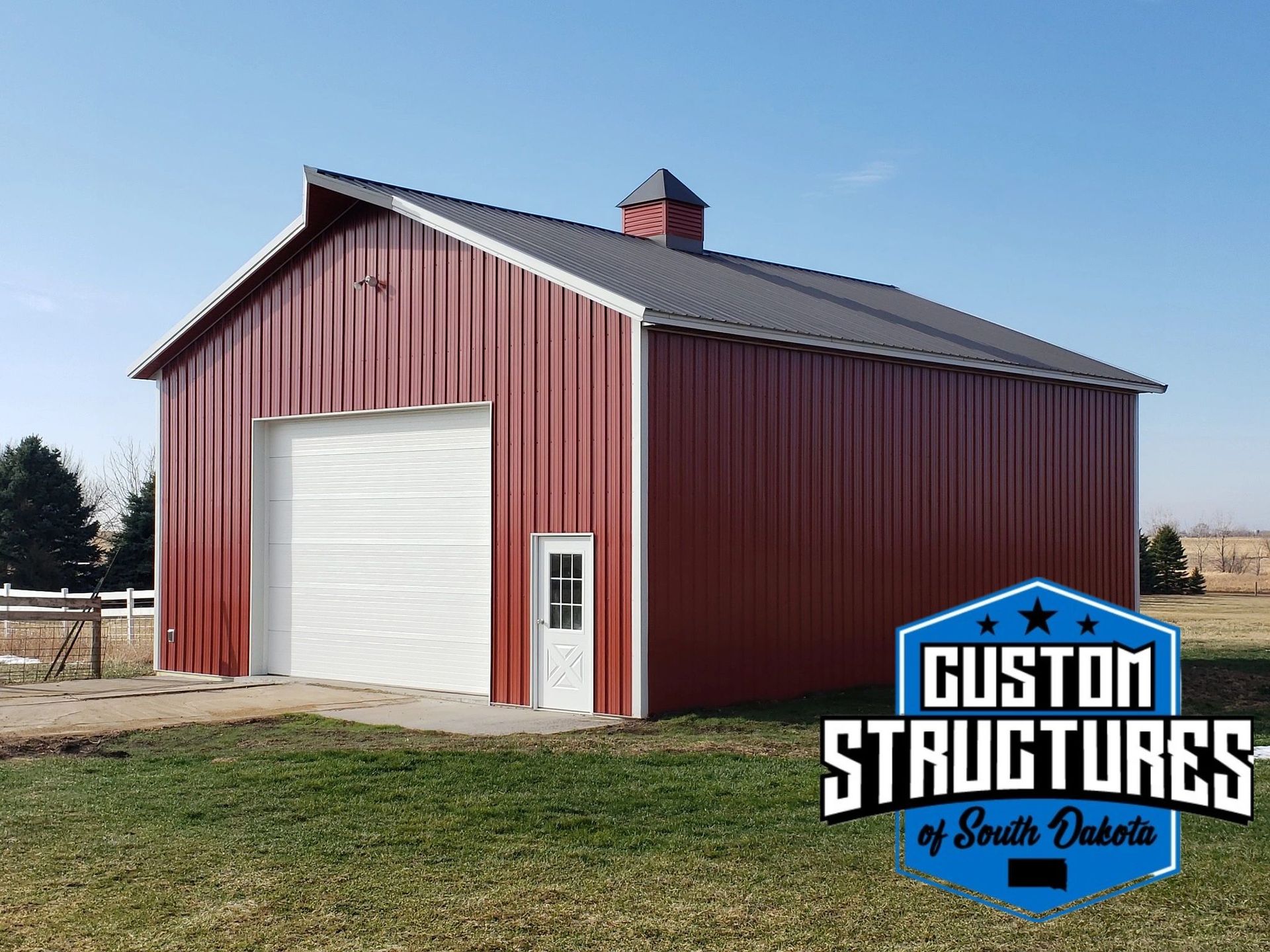 Red metal barn with white garage door and small door, logo of Guston Structures.