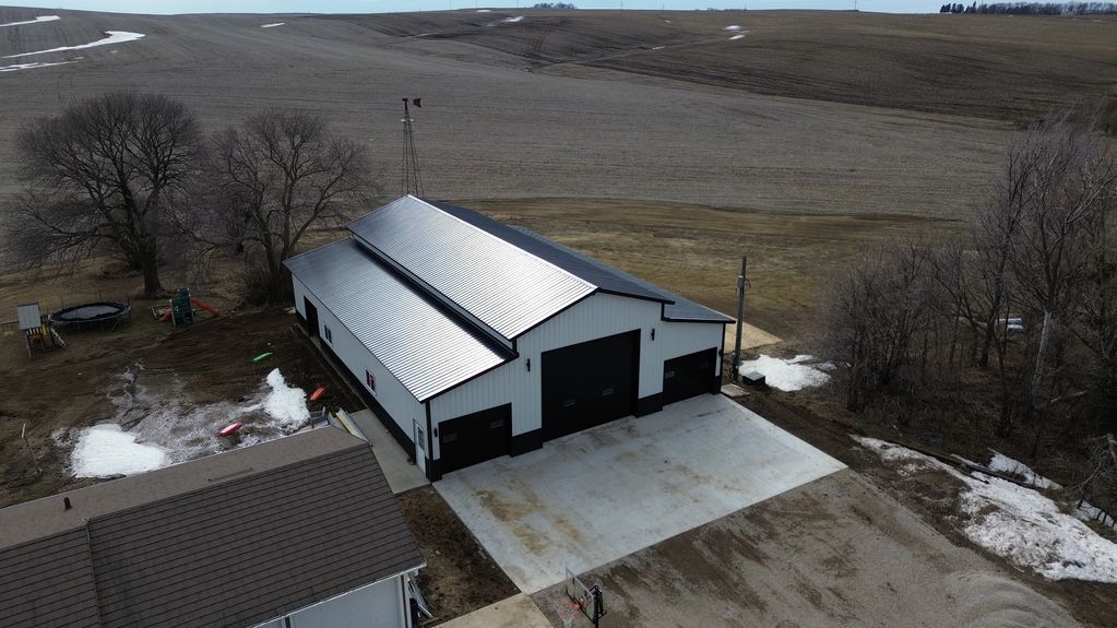 Aerial view of a white building with two garage doors and a metal roof on a concrete pad in a rural area.