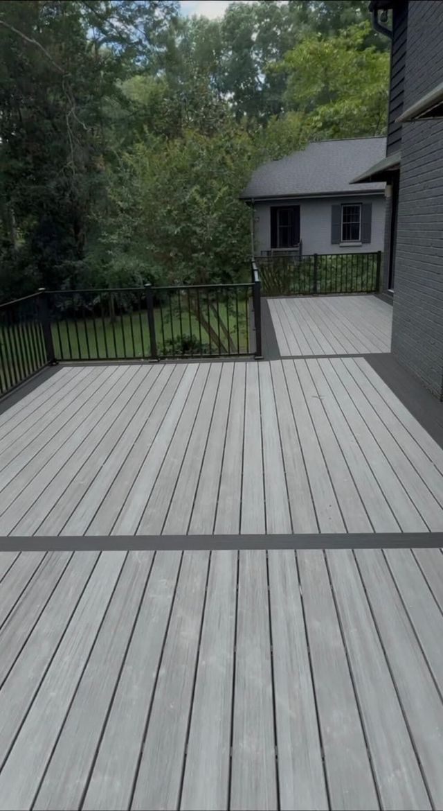 Gray wooden deck overlooking trees and a house, with black railing and walkway to a patio.