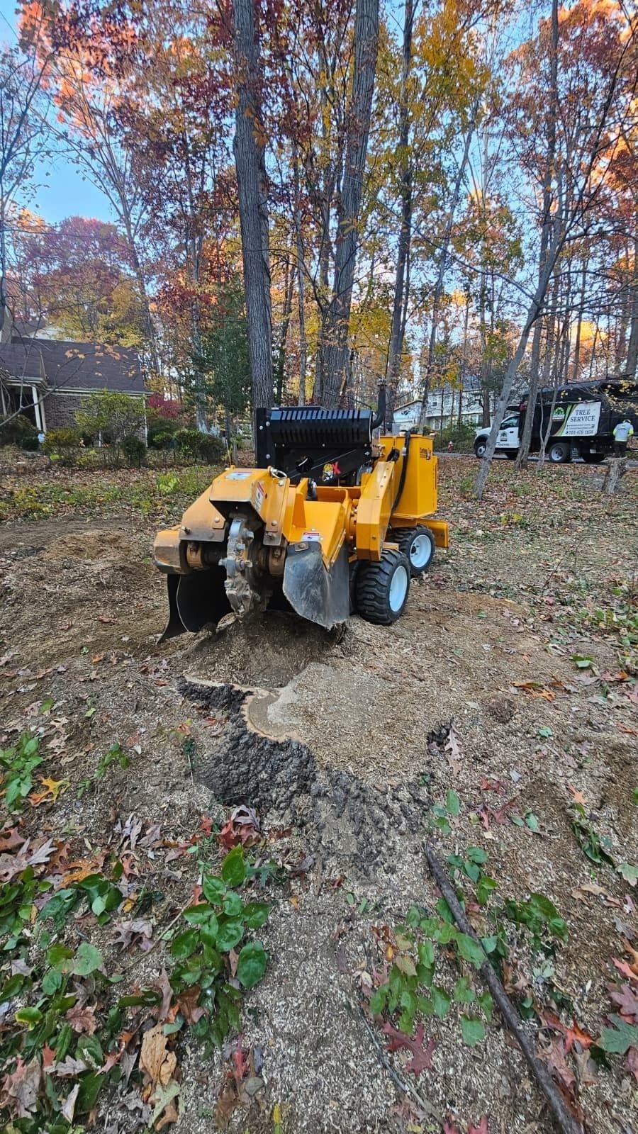 Yellow tractor attachment in a leaf-covered wooded yard, with trees and a truck in the background