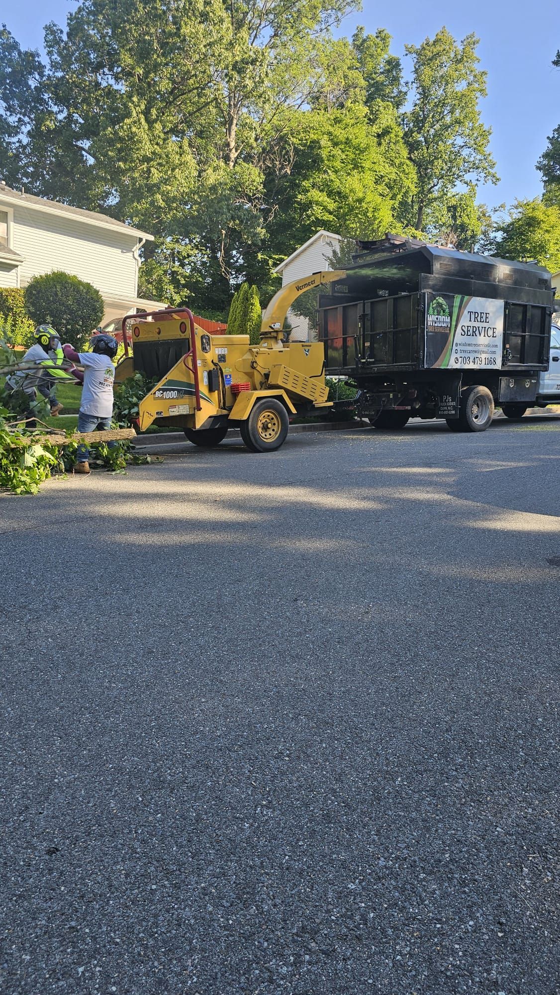 Workers loading yard debris into a truck with a yellow skid steer on a residential street.
