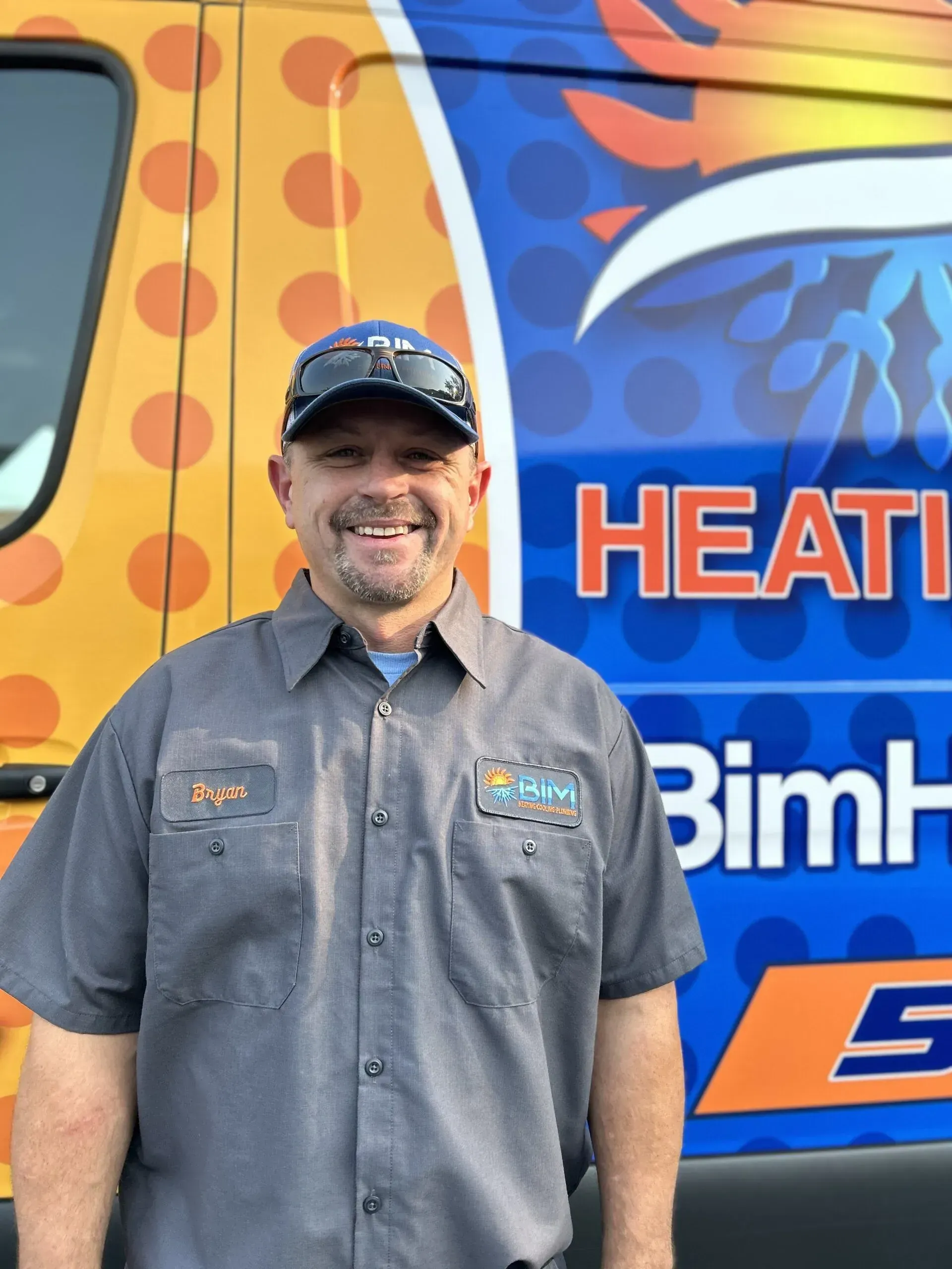 Smiling man in gray shirt and cap standing before a colorful HVAC service truck.