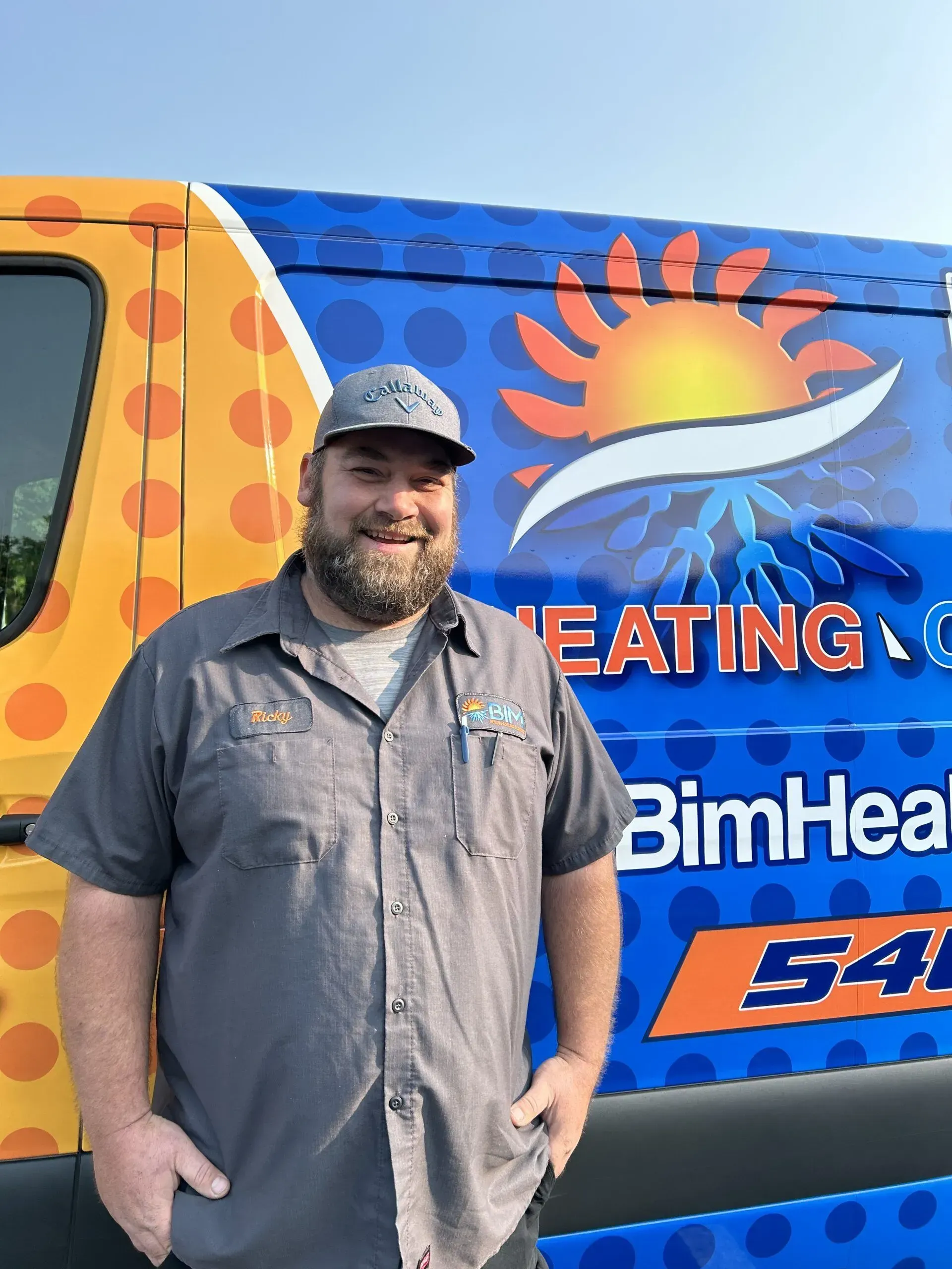 Smiling man in a gray shirt and cap standing beside a colorful HVAC service van