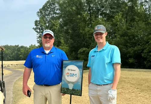 Two men in blue shirts stand beside a sign at a sunny outdoor golf course.