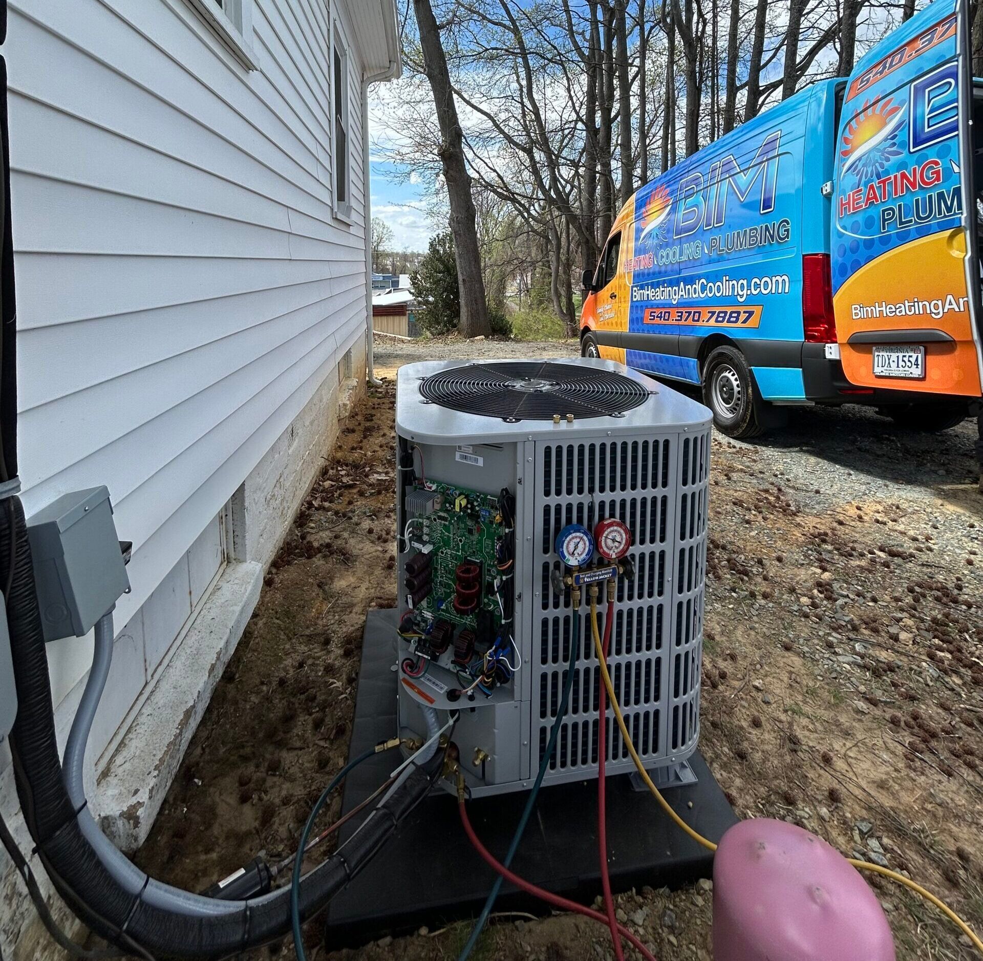 Outdoor HVAC unit beside a house, with service hoses connected and a colorful utility van nearby.