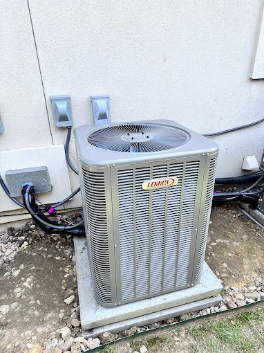 Outdoor air conditioning unit beside a building wall, with pipes and electrical boxes visible.