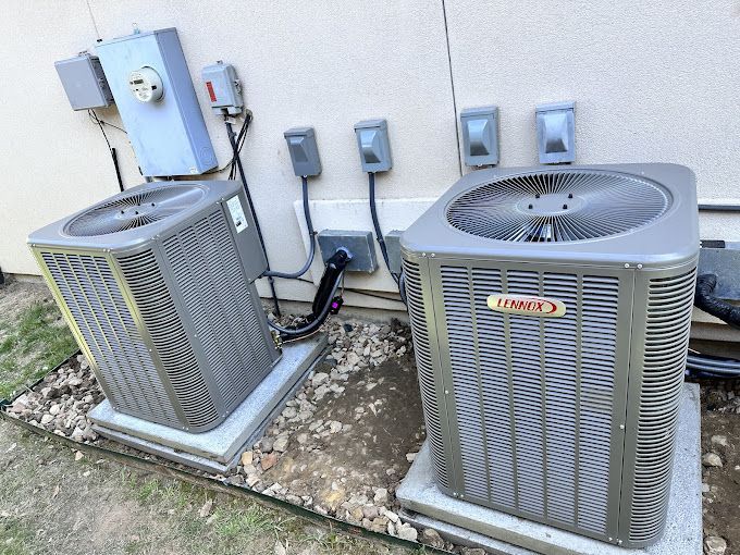 Two outdoor HVAC units beside a house wall, with electrical boxes and conduit behind them.