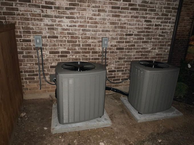 Two outdoor HVAC units on concrete pads against a brick wall with electrical conduit nearby