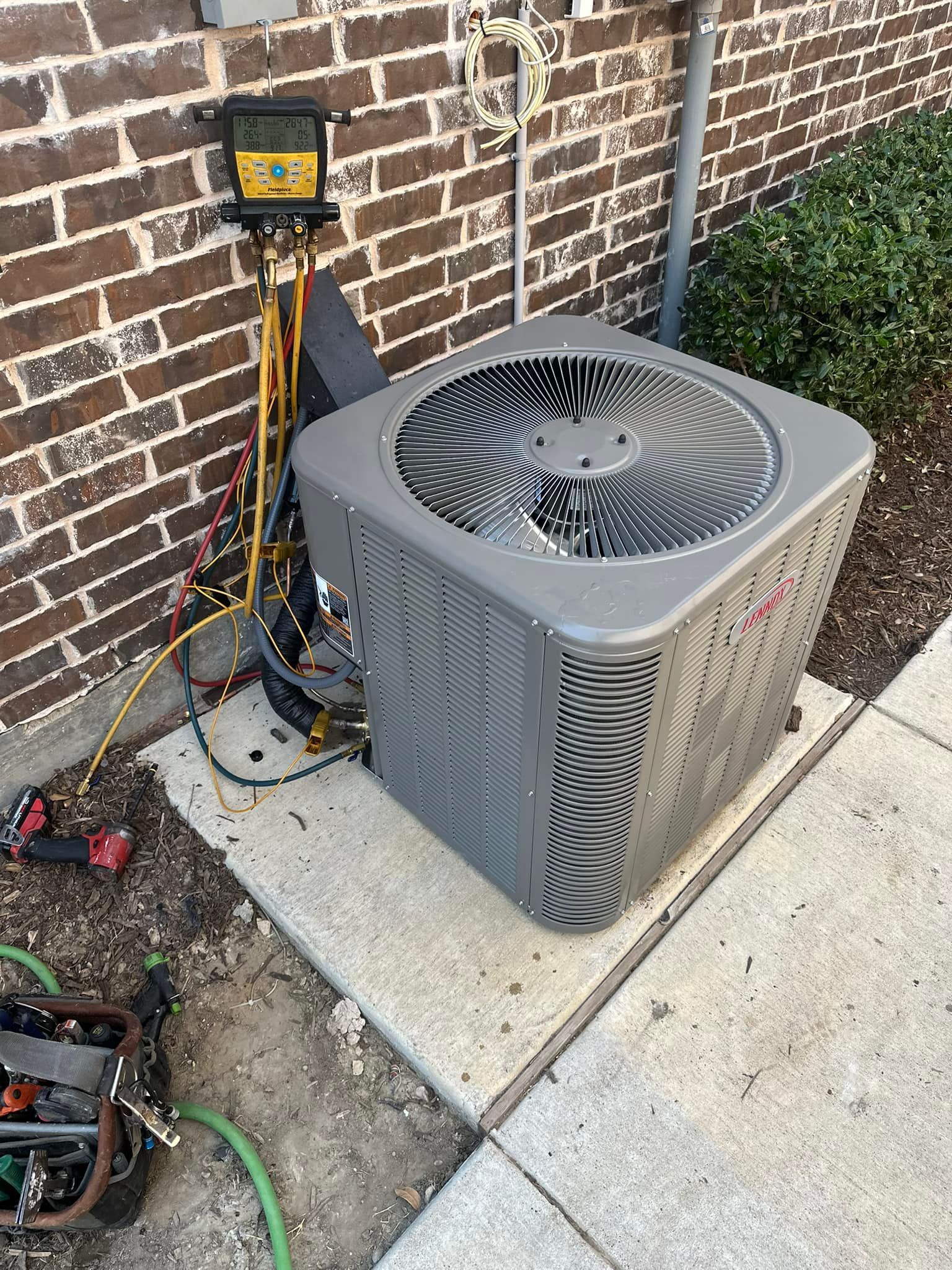 Outdoor air conditioner unit beside a brick wall, with a service gauge and hoses attached