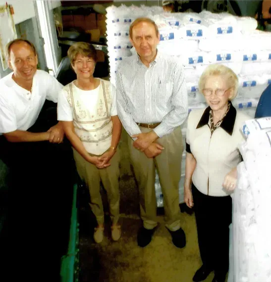 Four people standing in a storage area next to stacks of bagged ice.