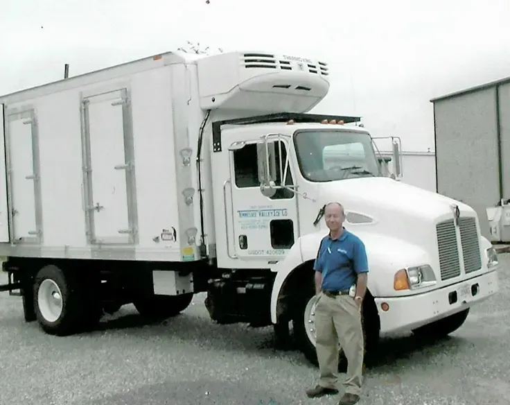 Man standing next to a white refrigerated truck. Gray pavement and building in the background.