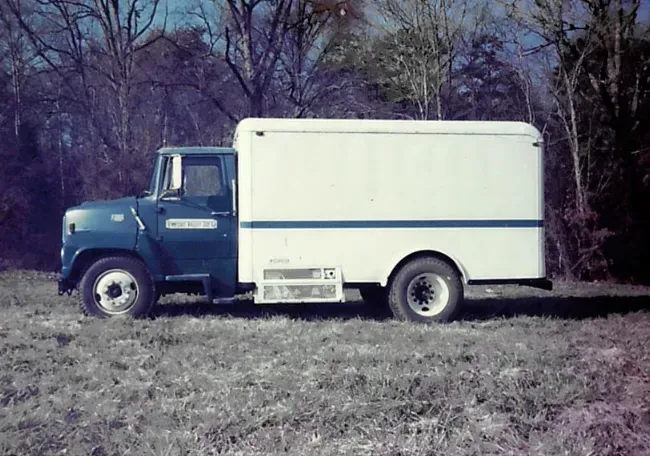 Blue and white International Harvester step van parked in a field with trees in the background.