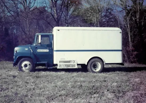 Blue and white International Harvester step van parked in a field with trees in the background.