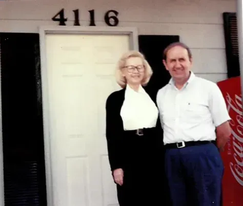 A man and woman stand in front of a door with the number 4116. A Coca-Cola machine is to the right.