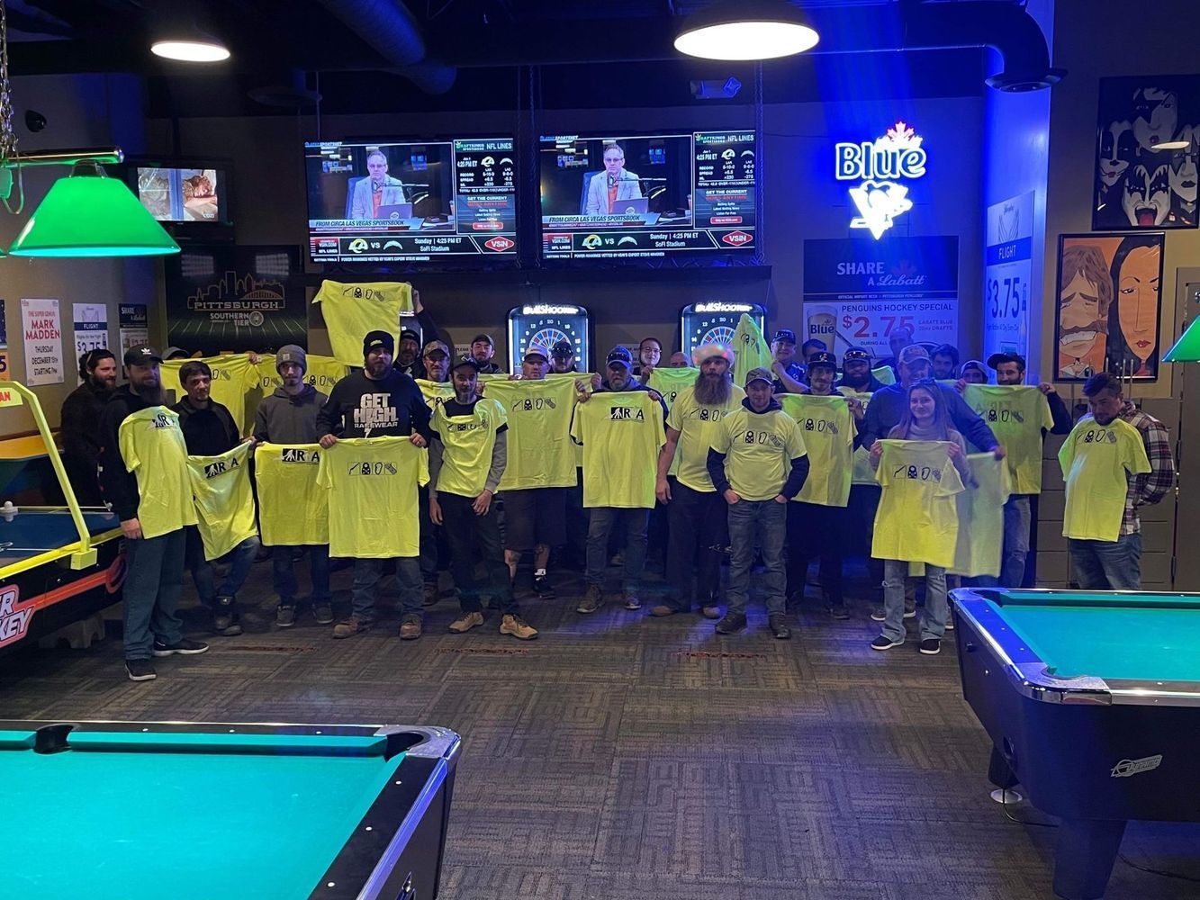 A group of people are standing in a pool hall holding yellow shirts.