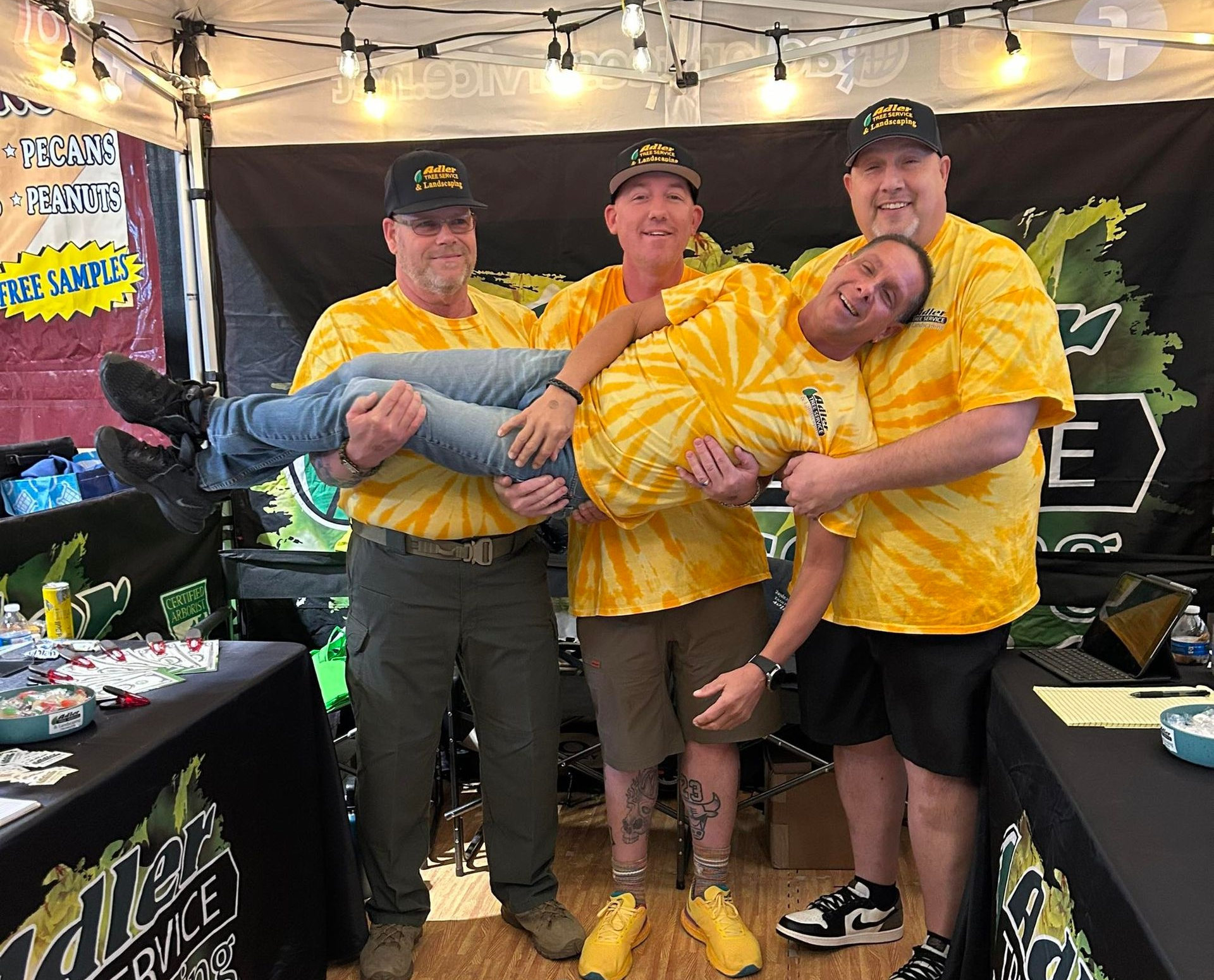Four men in yellow shirts and hats, one being held up, smiling, at a landscaping booth.