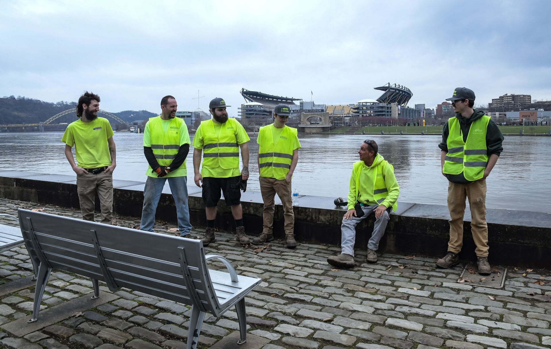Six people wearing lime-green safety vests stand or sit by a river near a stadium under a cloudy sky.