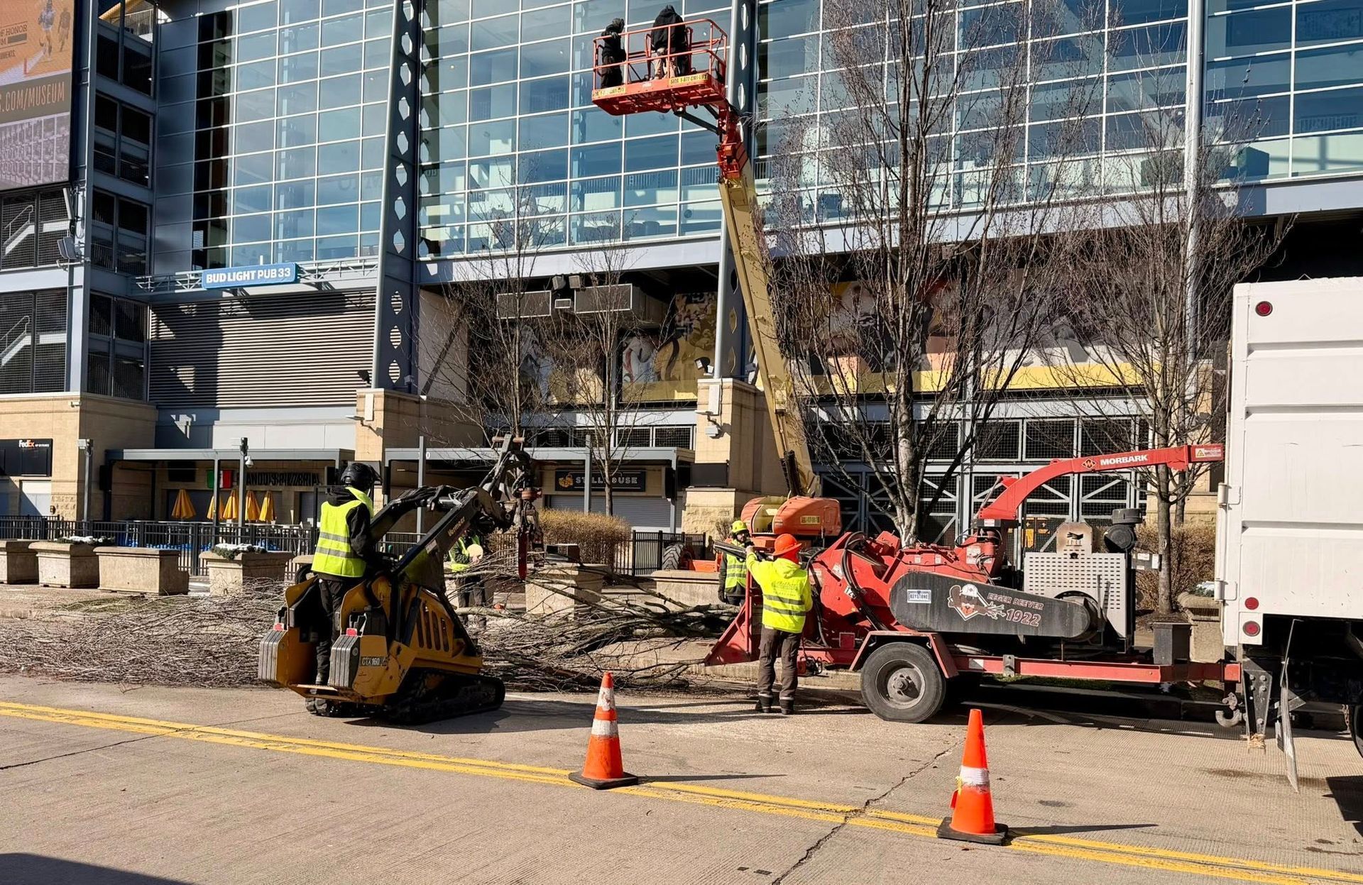 Workers use a yellow skid steer, a cherry picker, and a wood chipper for landscaping work in front of a modern building.