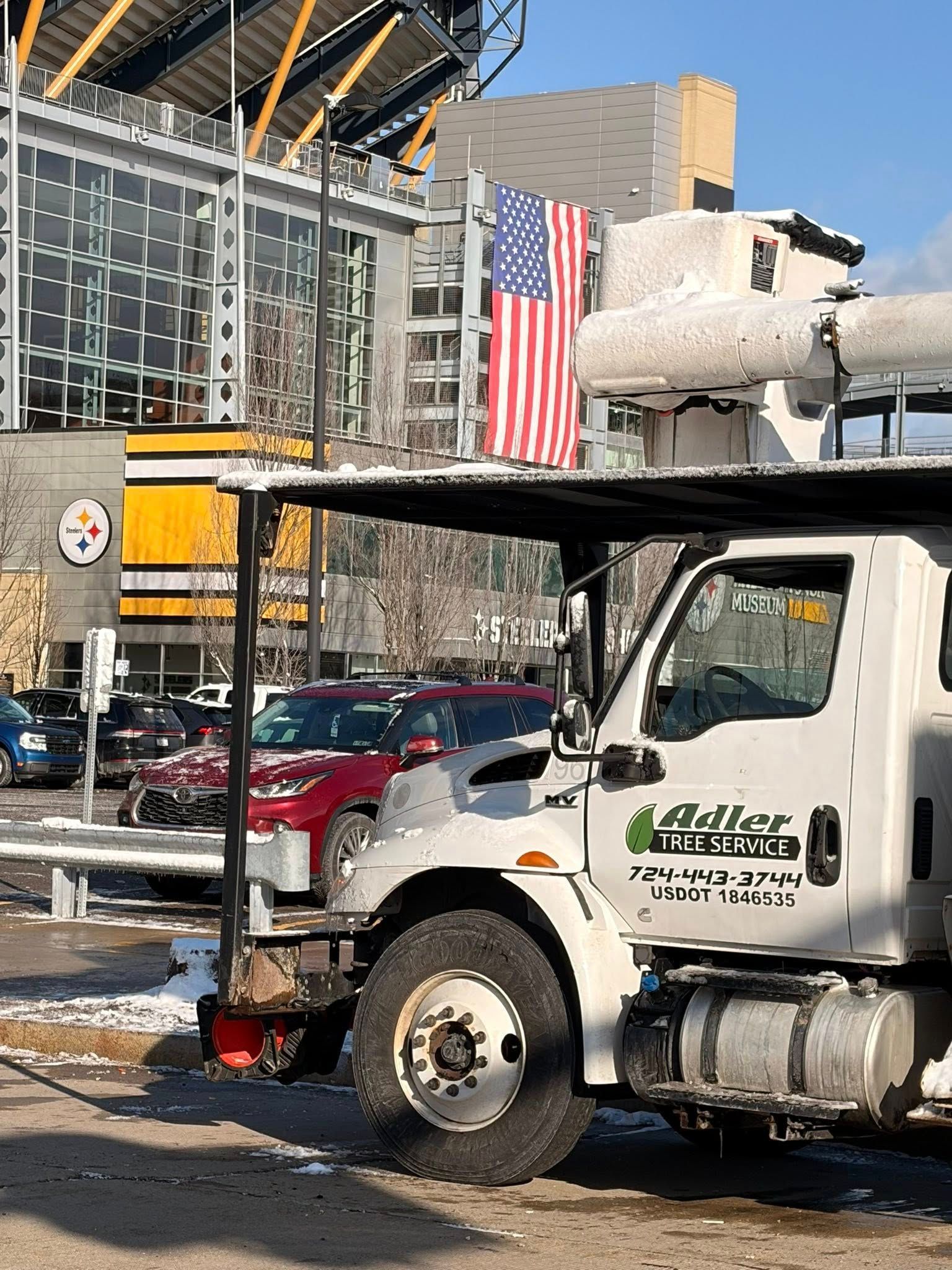 A white Adler utility truck is parked on a snowy day with an American flag displayed behind it in front of a stadium.