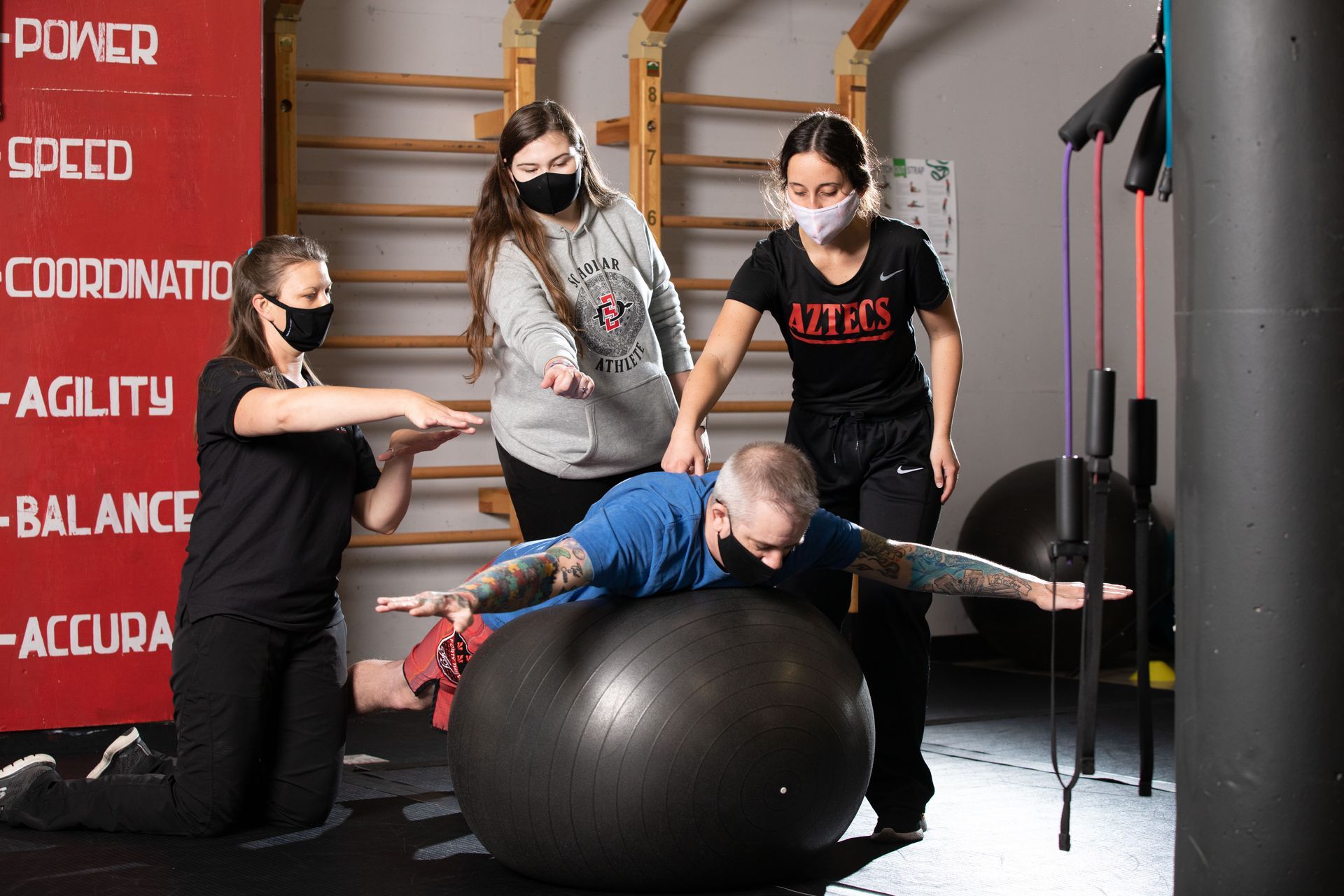 People exercising in a gym, with a child balancing on a black stability ball.