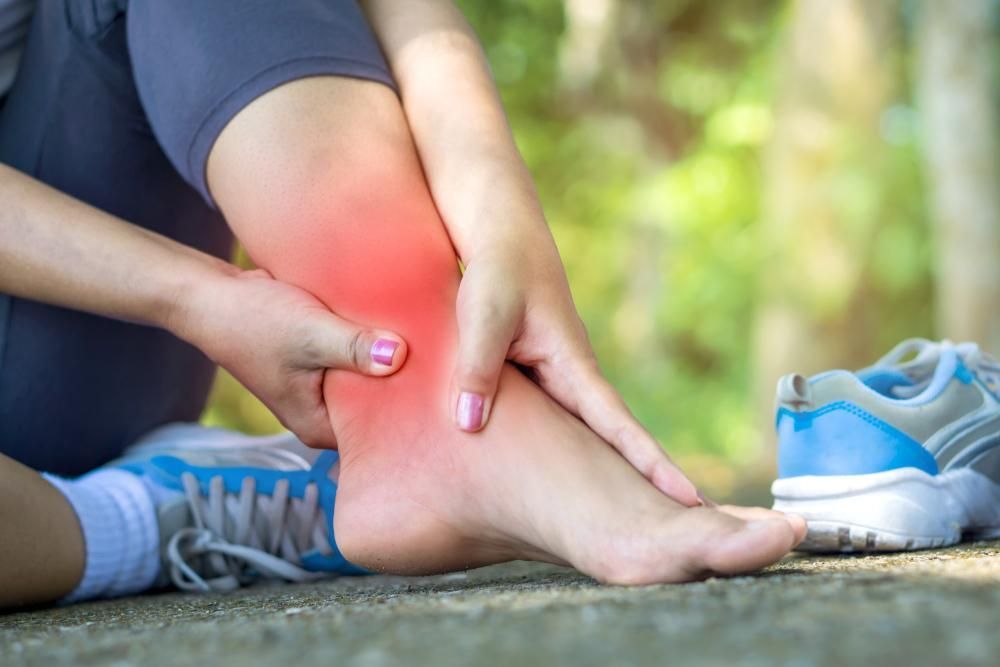 Person holding a red, painful ankle while sitting beside a blue sneaker outdoors
