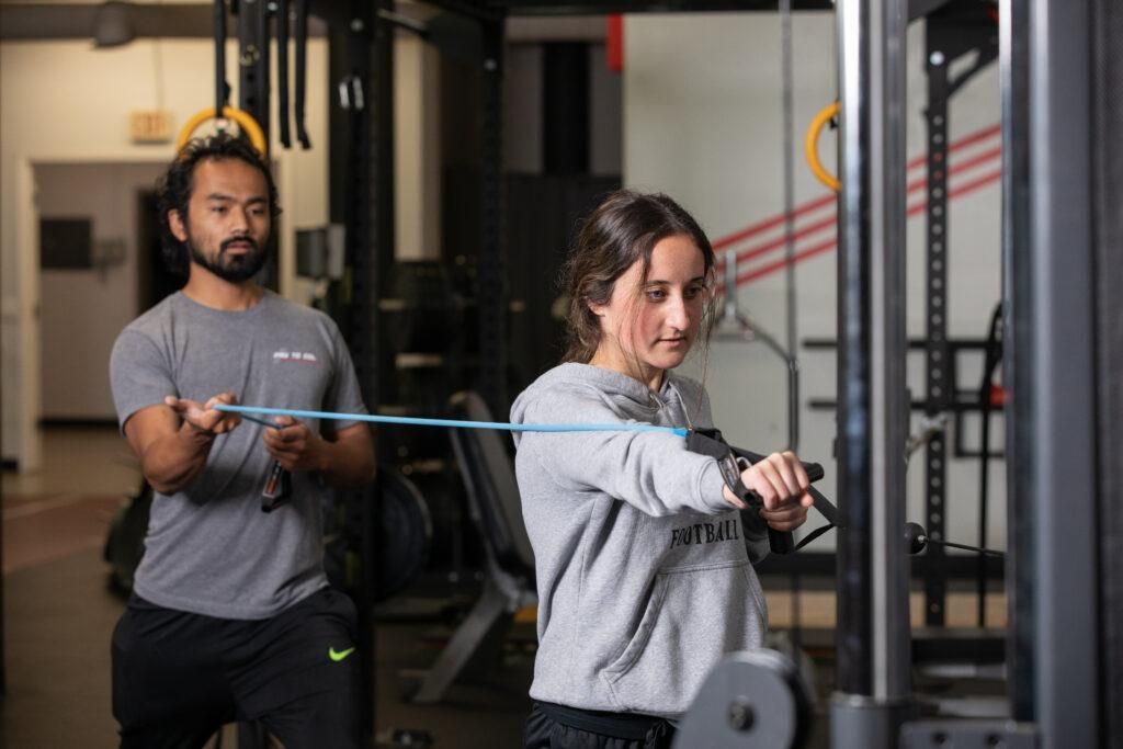 Two people doing cable exercises in a gym, one in front pulling a resistance band or cable.