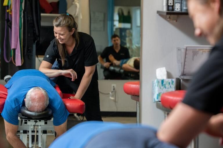 Massage therapist stretching a client's back in a clinic, with another client blurred in the foreground
