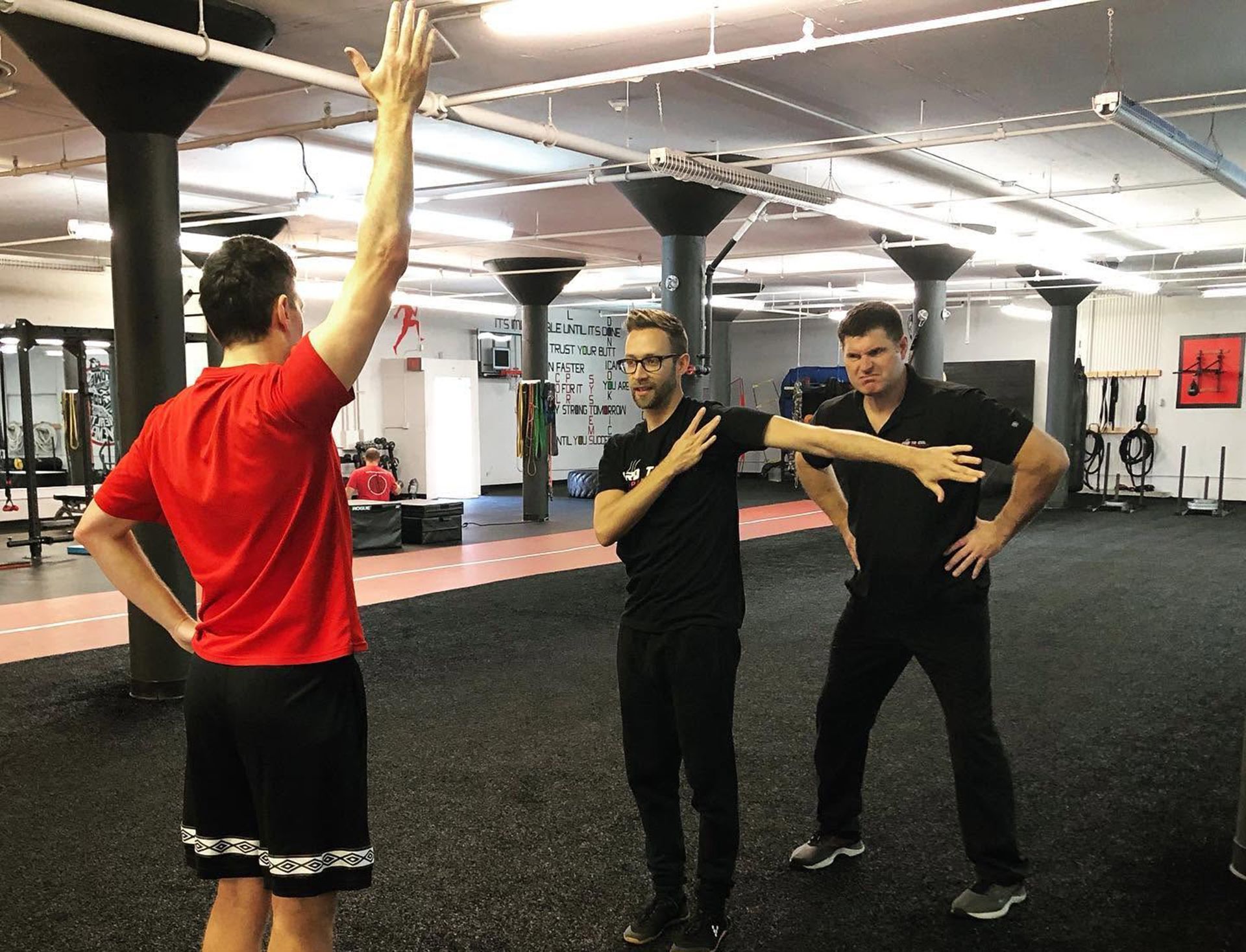 Three people in a gym practice a synchronized arm exercise on black flooring.