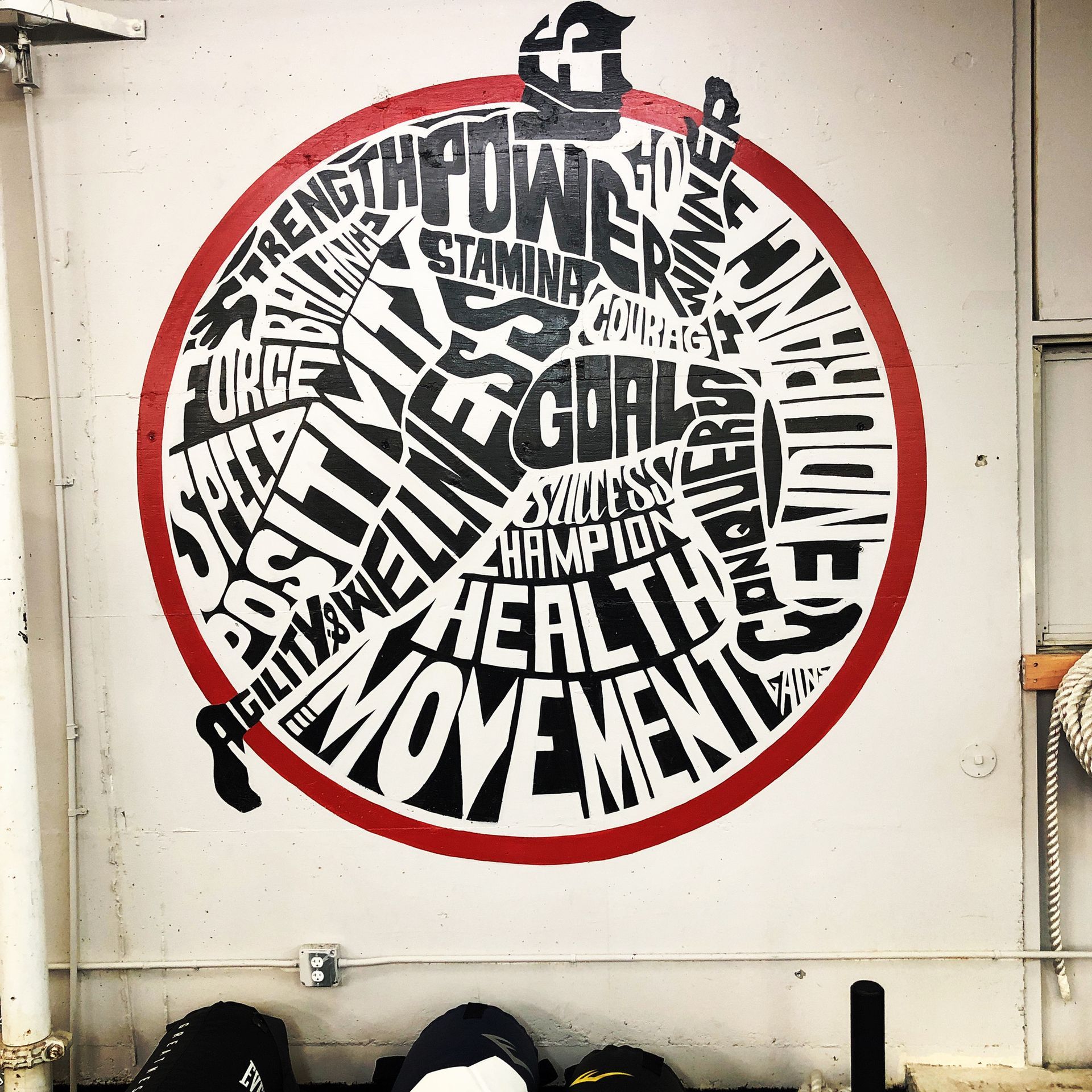 Circular black-and-red mural of overlapping fitness words on a white wall, with a small black figure on top.